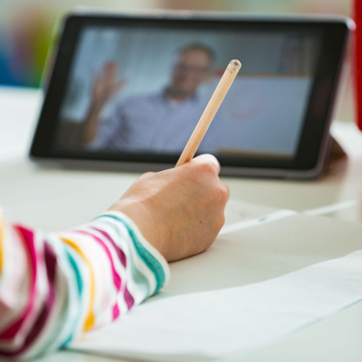 Eine junge Hand hält einen Stift und schreibt damit auf Papier. Im Hintergrund steht auch ein Tablet auf dem Tisch, auf dem eine Lehrkraft verschwommen zu sehen ist.