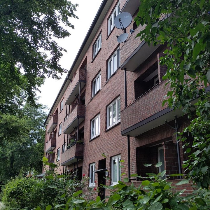 Ein Mehrparteienhaus mit Balkone. Der rote Backstein Sticht hervor. Drum herum: Grüne Bäume und Pflanzen.