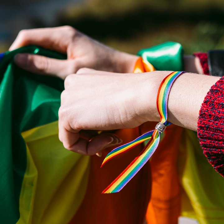 Eine Regenbogenfahne wird gehalten. Die Hände dazu haben ein Regenbogenbändchen.